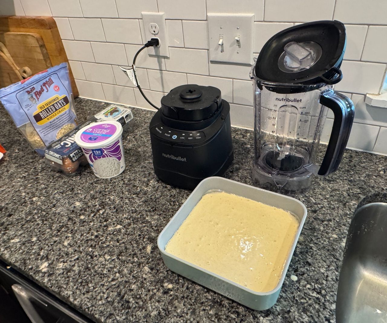 Batter inside a square cake tin besides a cleaned out blender pitcher on a stone kitchen worktop.