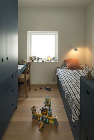 Interior view of a bedroom at the Oslo family house featuring white walls, wood flooring, a window, a blue wardrobe, drawers and desk with a wooden chair, a blue bed with striped linen and a cushion, a white wall lamp and toys on the window sill and floor