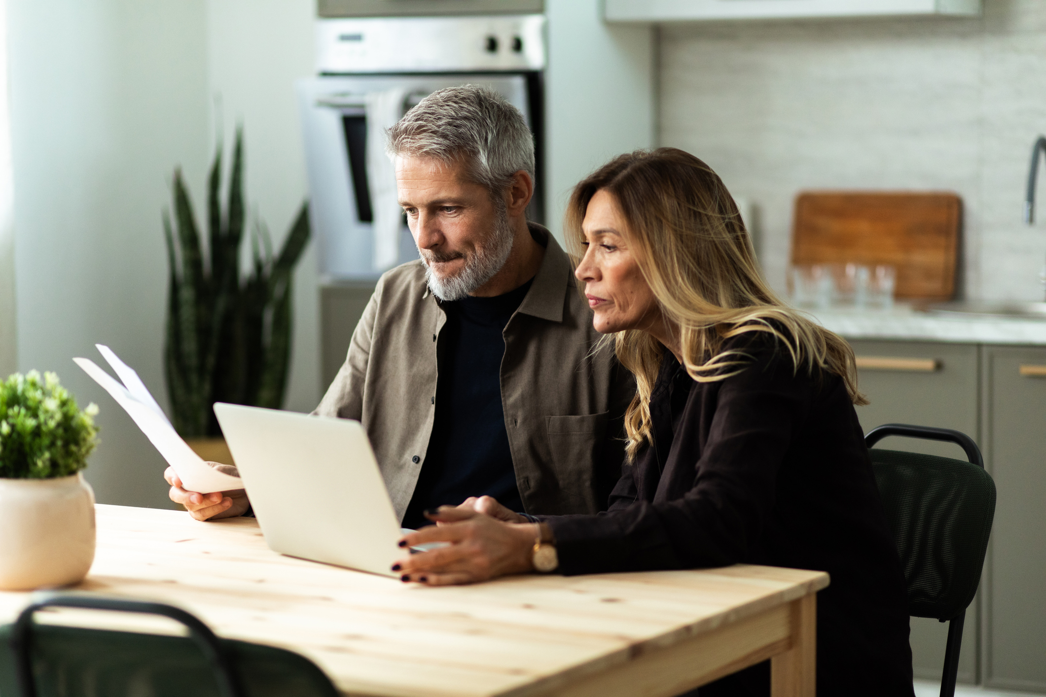 Mature couple discussing finances at home, looking at laptop and holding paperwork.