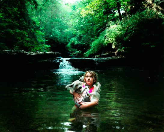 A little girl carrying a dog through a lake with the forest behind them.