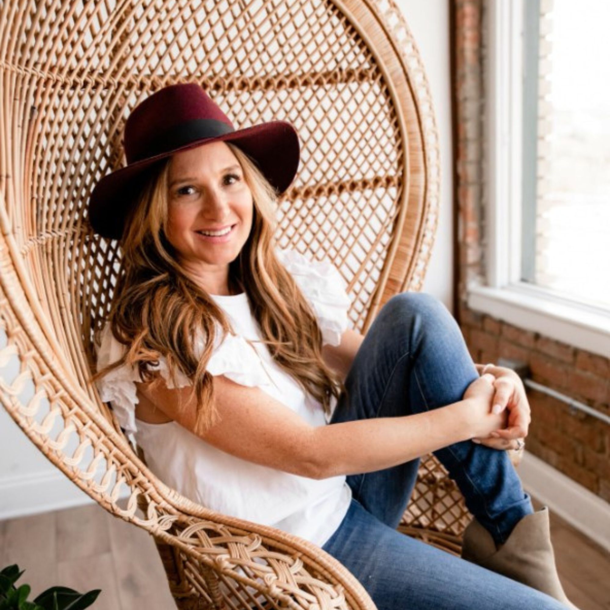 A picture of Daniella Menachemson in a white t-shirt, jeans, and a hat, sitting in a wicker chair