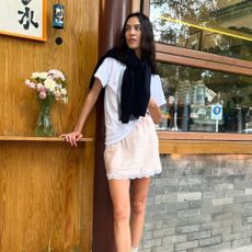 Alexa Chung stands outside wearing a pink satin miniskirt with lace-trim detailing with black trainers, a white t-shirt and a jumper wrapped around her shoulders.