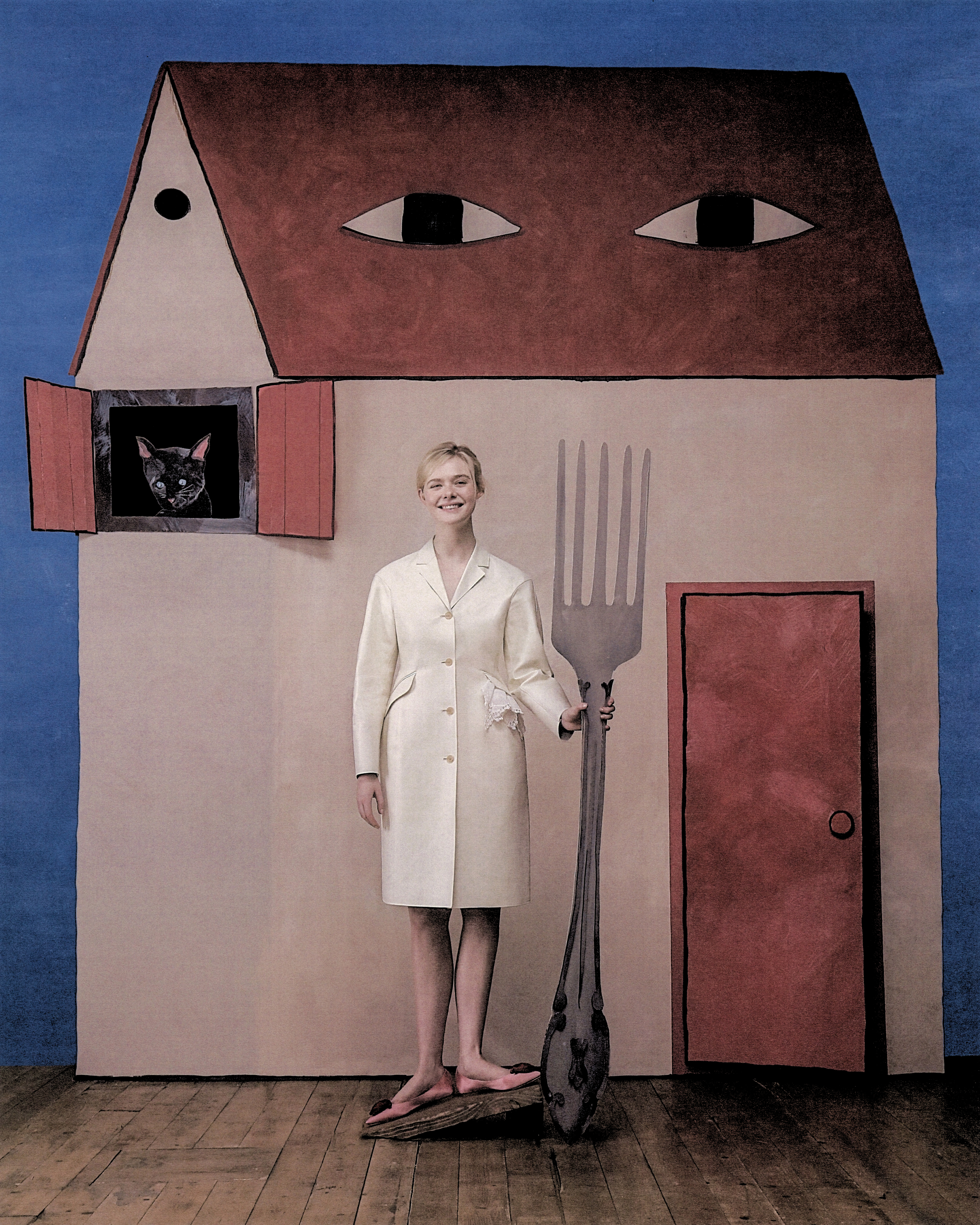 Elle Fanning shot by Szilveszter Mak&amp;oacute;. Fanning is seen standing in front of a large cardboard cutout house with a cardboard black cat peeking out one the window. She is wearing a white buttoned coat and holding an oversize cardboard cutout of a fork.