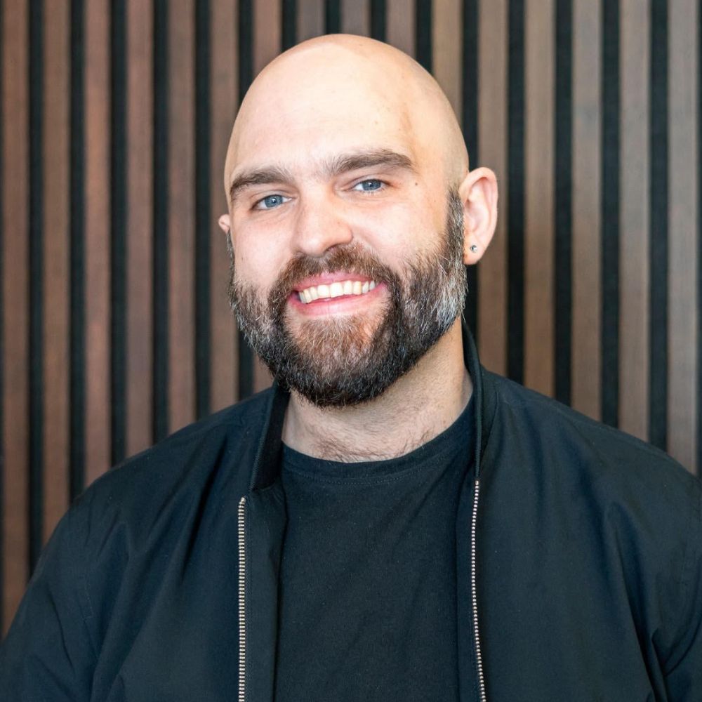 A smiling man with a beard standing against a slatted wood wall