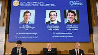 Three men sit at a table to announce the Nobel Prize in chemistry, with the winners on a screen behind them