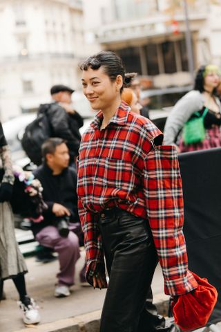 A woman at fashion week wearing a flannel shirt outfit of a red tartan shirt, leather pants, and a red clutch purse.