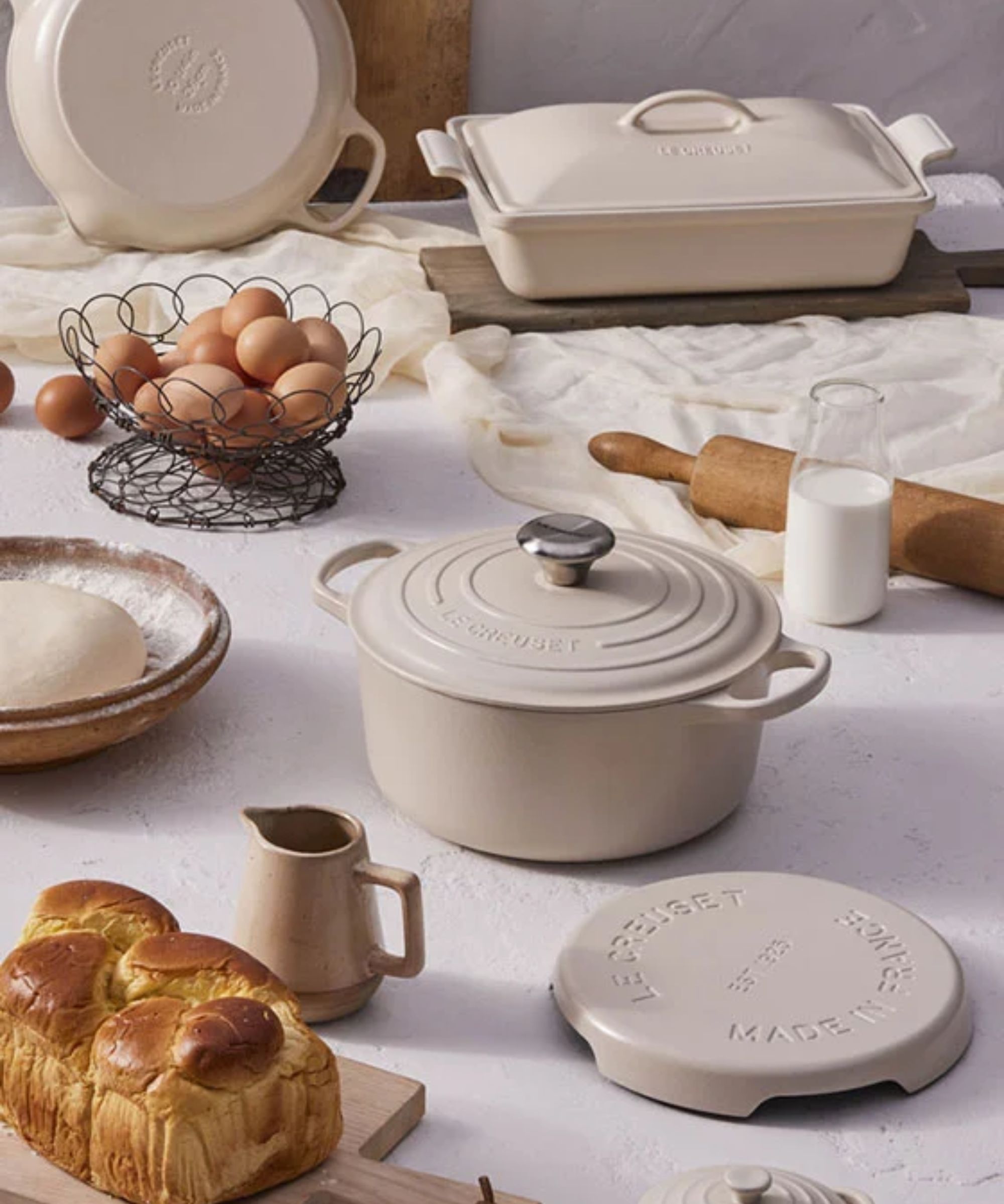 The Le Creuset cast iron cookware in the Brioche colorway, over a white table, with brioche bread and a dish of eggs.