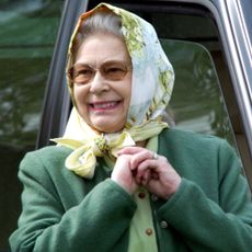 Queen Elizabeth wearing a head scarf and a green coat smiling in front of a car