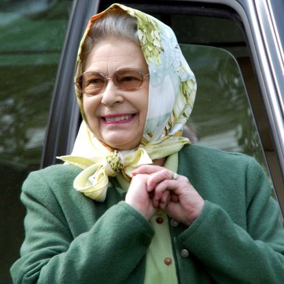 Queen Elizabeth wearing a head scarf and a green coat smiling in front of a car