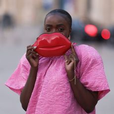 a woman wearing a hot pink dress holds her lip-shaped handbag over her mouth