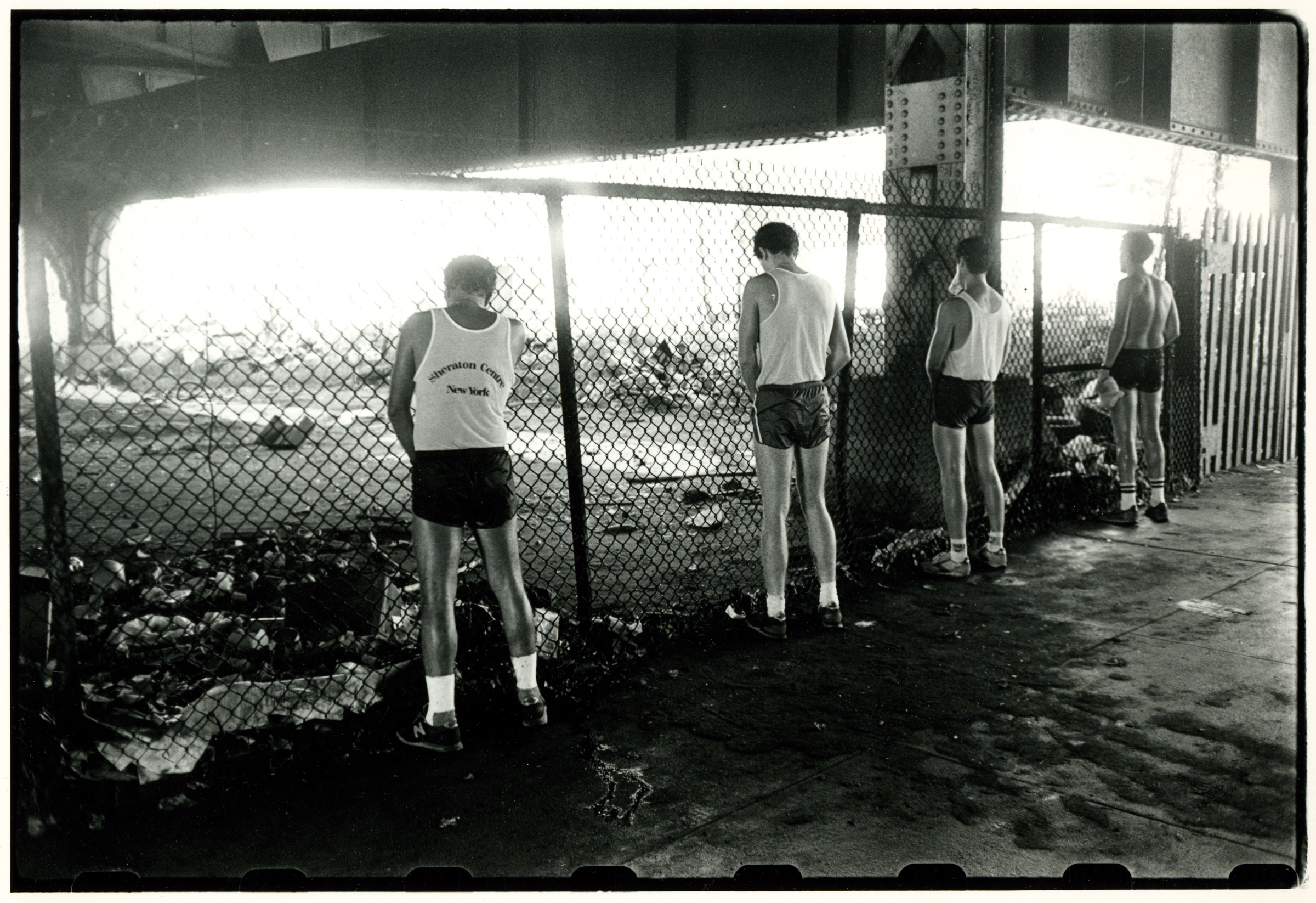 Four runners in vests and shorts stand with their backs to the camera against a chain-link fence beneath an overpass
