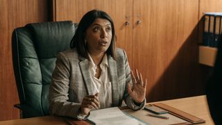 DCI Maiya Goswami (Sunetra Sarker) sits in a high-backed leather chair at her desk, holding up her hands in some sort of protesting gesture