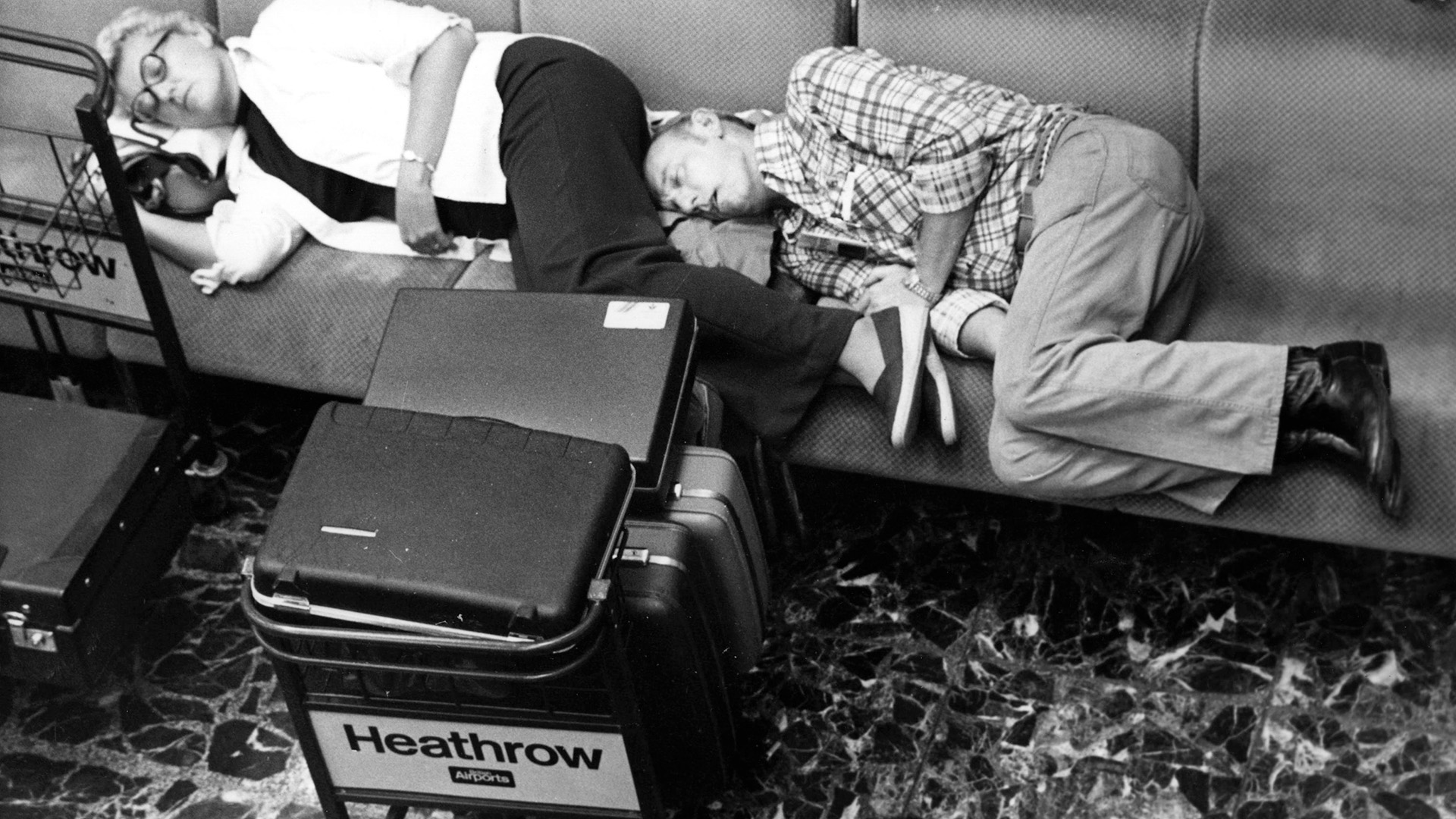 A black and white photo of two people sleeping on a bench seat with suitcases next to them labeled "Heathrow"