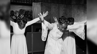 Black and white photograph of three women, wearing white dresses. They have their heads bent down with their arms outstretched.