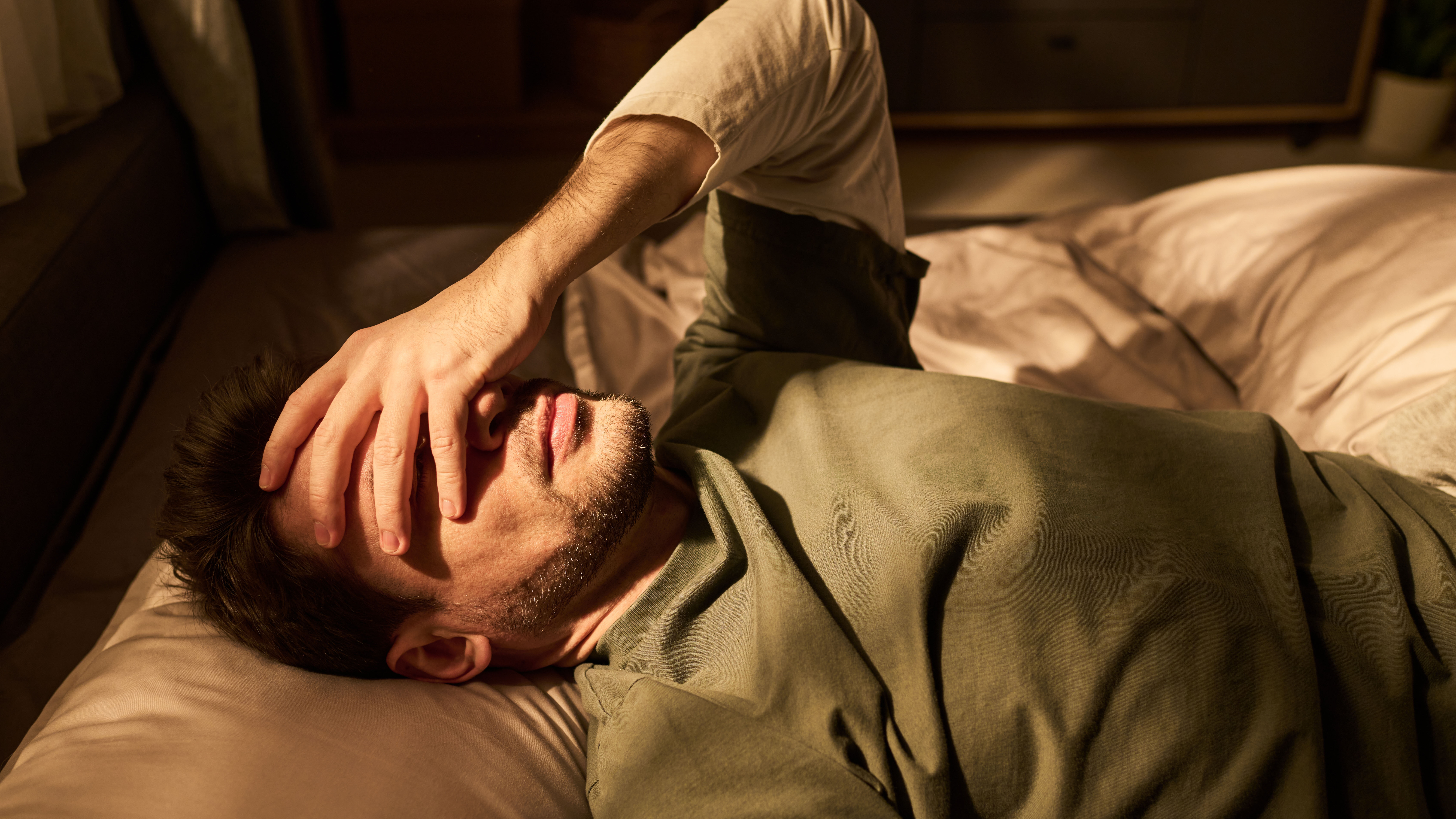 Man lying on bed covering face with hand appearing stressed or exhausted.