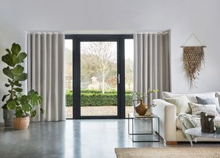 A neutral living room with large French windows leading to a garden with long white curtains