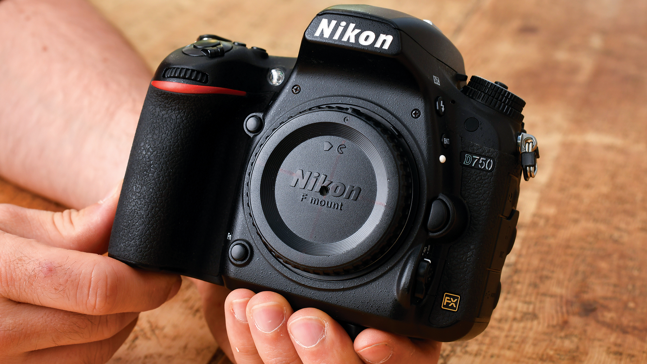 Close-up of a Nikon D750 camera held in hand, showcasing the F mount and textured grip against a wooden surface