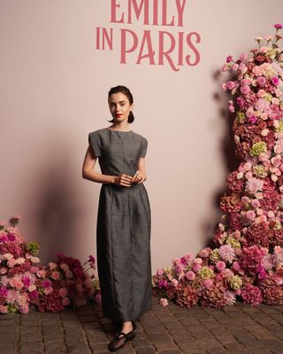 Lily Collins stands in front of a pink wall with rose detailing wearing a gray mark kenly domino tan dress and alaia ballet flats