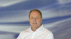 Dr Dave Smith, the new UK National Technology Adviser, sat in a white shirt against a background of solid blue broken up with clouds and white cross marks.