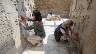 two women excavate cuneiform walls in narrow outdoor hallway of urartu temple