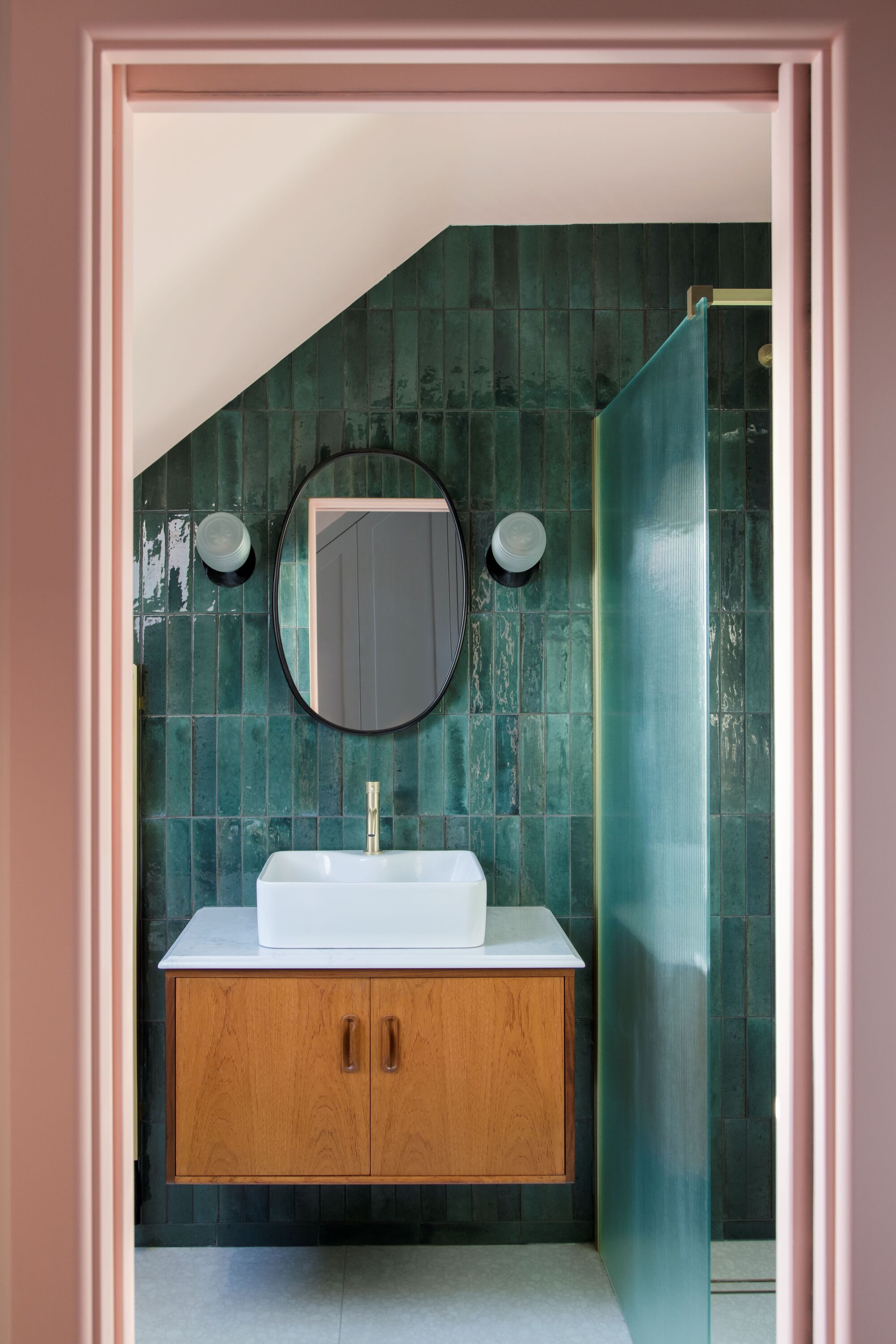 mid-century modern bathroom with green glossy subway tiles, a floating timber vanity with white sink and black round mirror with two wall sconces on each side, and angled roof, and pink walls outside the bathroom