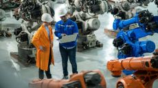Two factory workers wearing an orange and a blue jacket, looking at a laptop while surrounded by robotic arms for automated manufacturing.