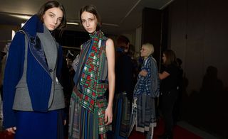 One woman in a blue and grey jacket and blue skirt, and one woman in a layered dress with tartan plaid design