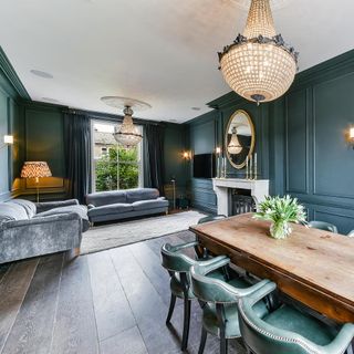 living and dining area with dark green walls, dark wooden floorboards, grey velvet sofas, a wooden dining table with green leather chairs, a fireplace with a white mantle and two gold feature pendant light fixtures