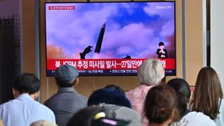 people watch a large tv screen with an image of a missile launching into a blue sky