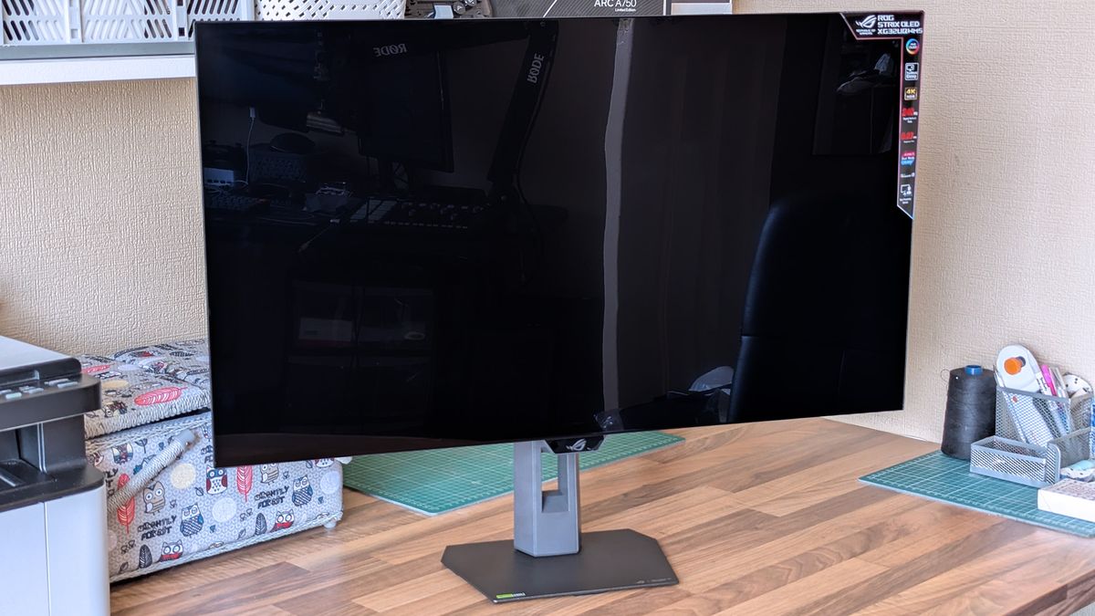 ASUS ROG Strix OLED monitor on a workspace desk, powered off, with crafting tools, organizers, and a second display visible in the background.