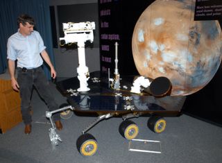 man stands next to a large rover module and looks down. at right is a poster of mars