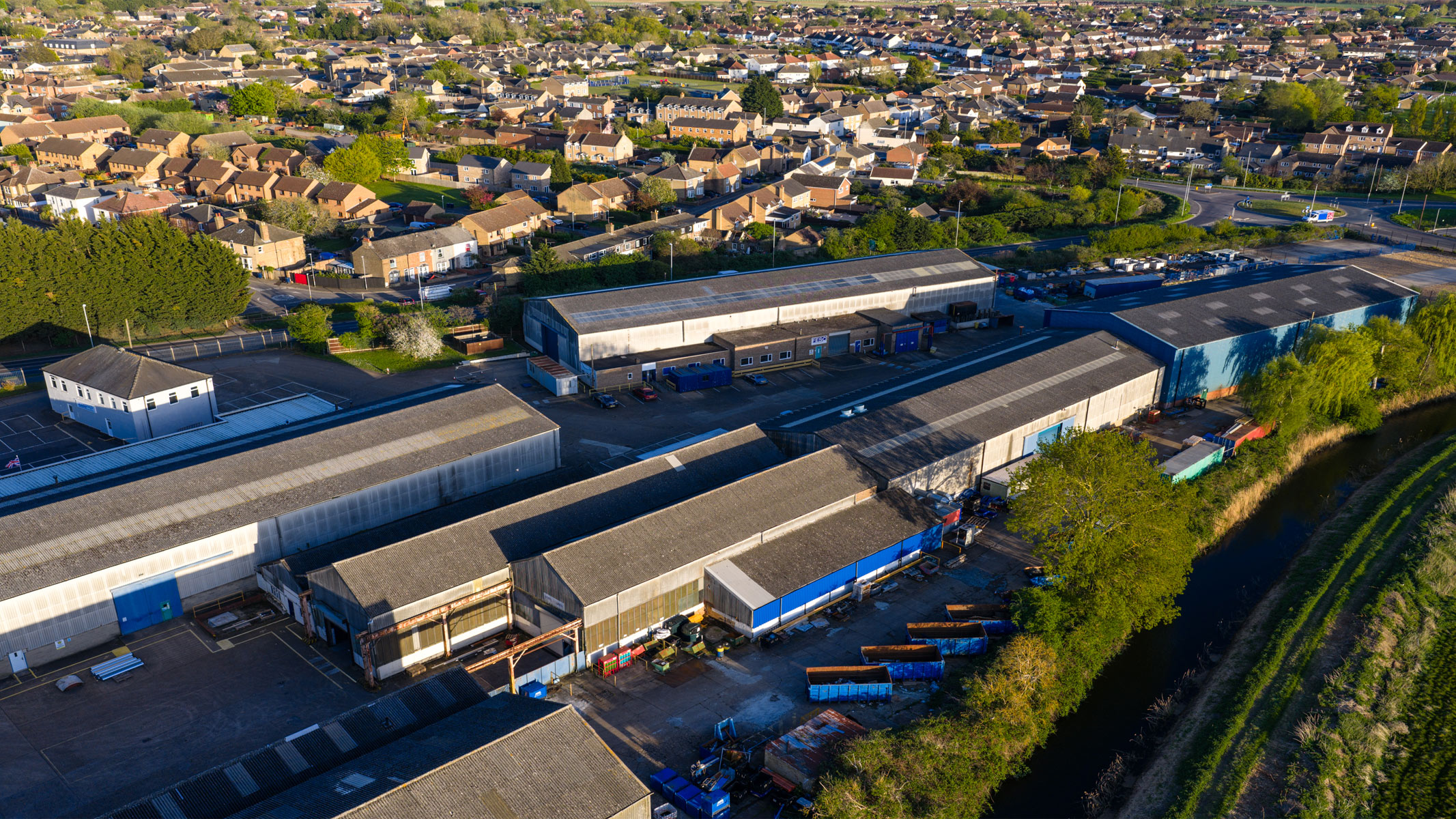 A bird's eye view of some depot/storage buildings next to a background of houses and some trees in between.