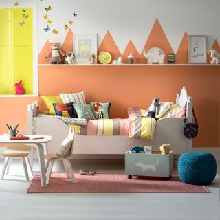 A childrens bedroom with a peach mountain graphic on the wall and a wooden bed, table and chairs and toy box