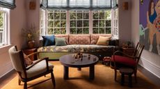 A living room with lilac walls, a bay window with blue Roman blinds, a sofa in the window with a floral seat cushions and green, rust, and dark blue scatter cushions. An ochre rug, modern coffee table and accent chair.