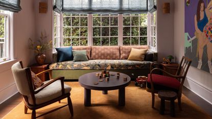 A living room with lilac walls, a bay window with blue Roman blinds, a sofa in the window with a floral seat cushions and green, rust, and dark blue scatter cushions. An ochre rug, modern coffee table and accent chair.