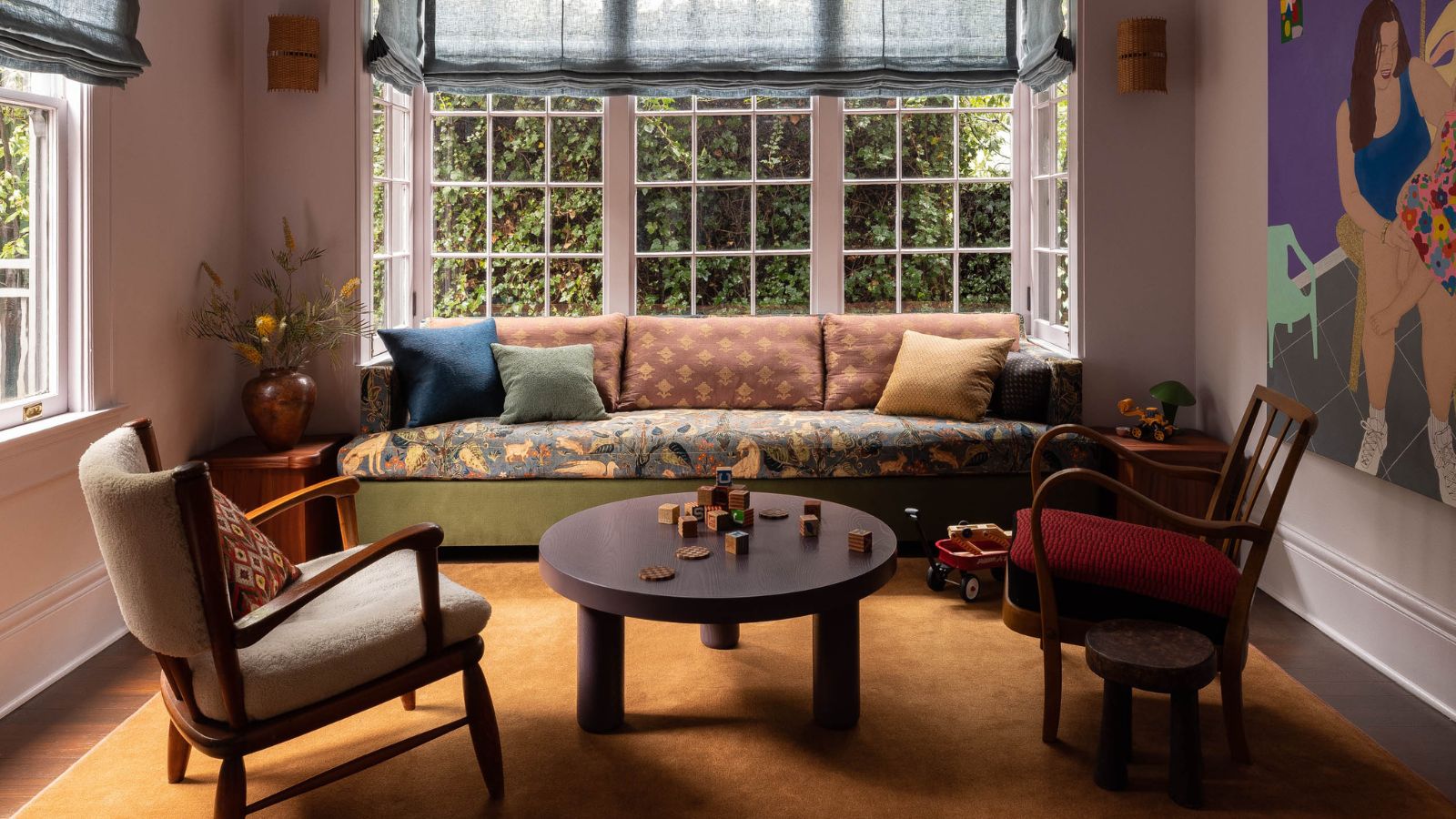 A living room with lilac walls, a bay window with blue Roman blinds, a sofa in the window with a floral seat cushions and green, rust, and dark blue scatter cushions. An ochre rug, modern coffee table and accent chair.