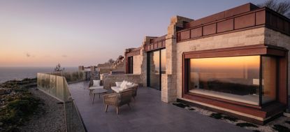 A terrace at sunset with rattan outdoor furniture and the sunset reflected in the large glass windows of the apartment