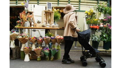 A woman wearing a hat and looking at a flower stall is pushing the Red Kite Baby Push Me 2U travel stroller