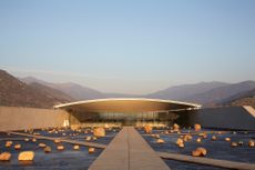 a low, horizontal building designed by Smiljan Radic, lit by orange sunlight sits among mountains, with a long path leading up to it, surrounded by rocks