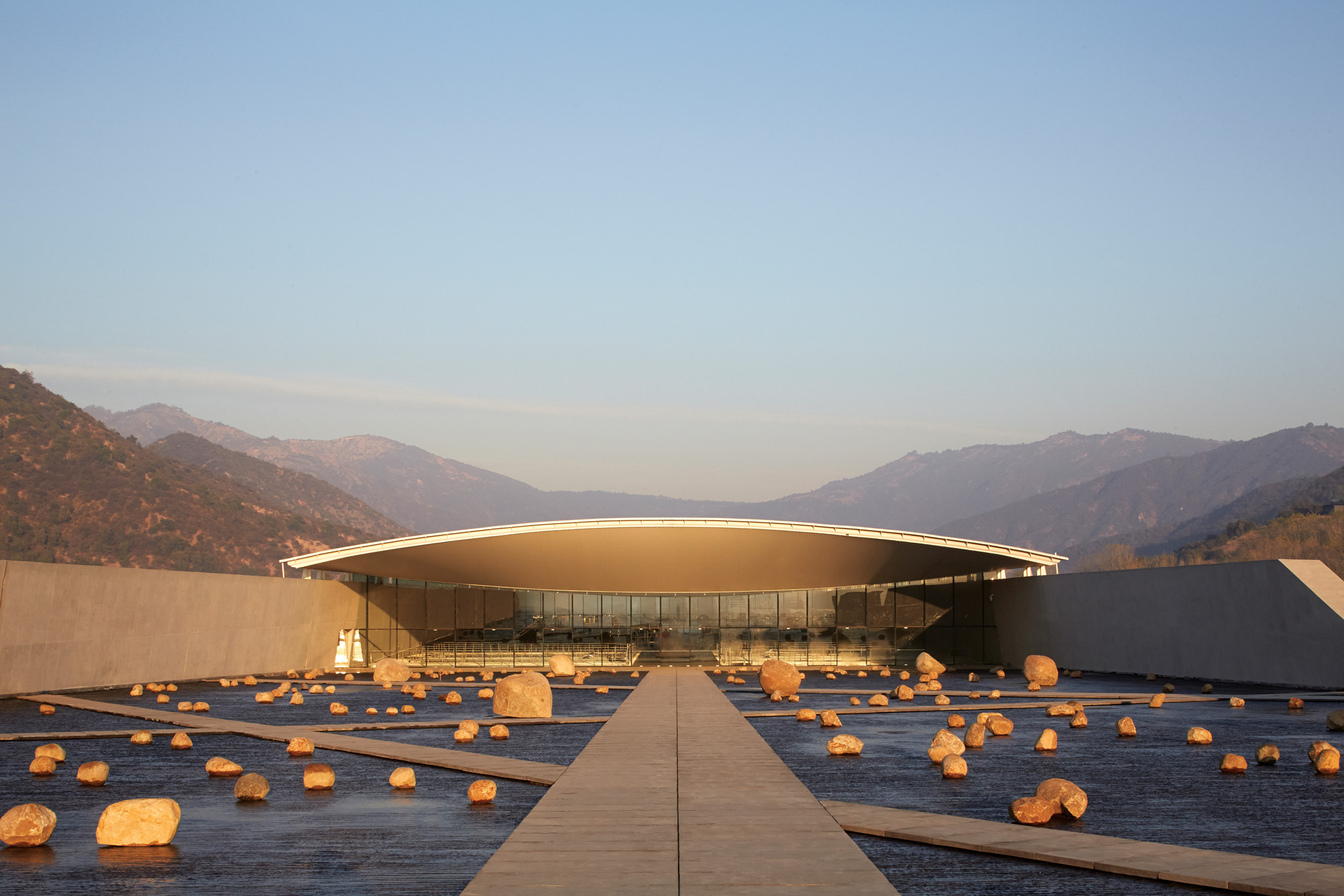a low, horizontal building designed by Smiljan Radic, lit by orange sunlight sits among mountains, with a long path leading up to it, surrounded by rocks