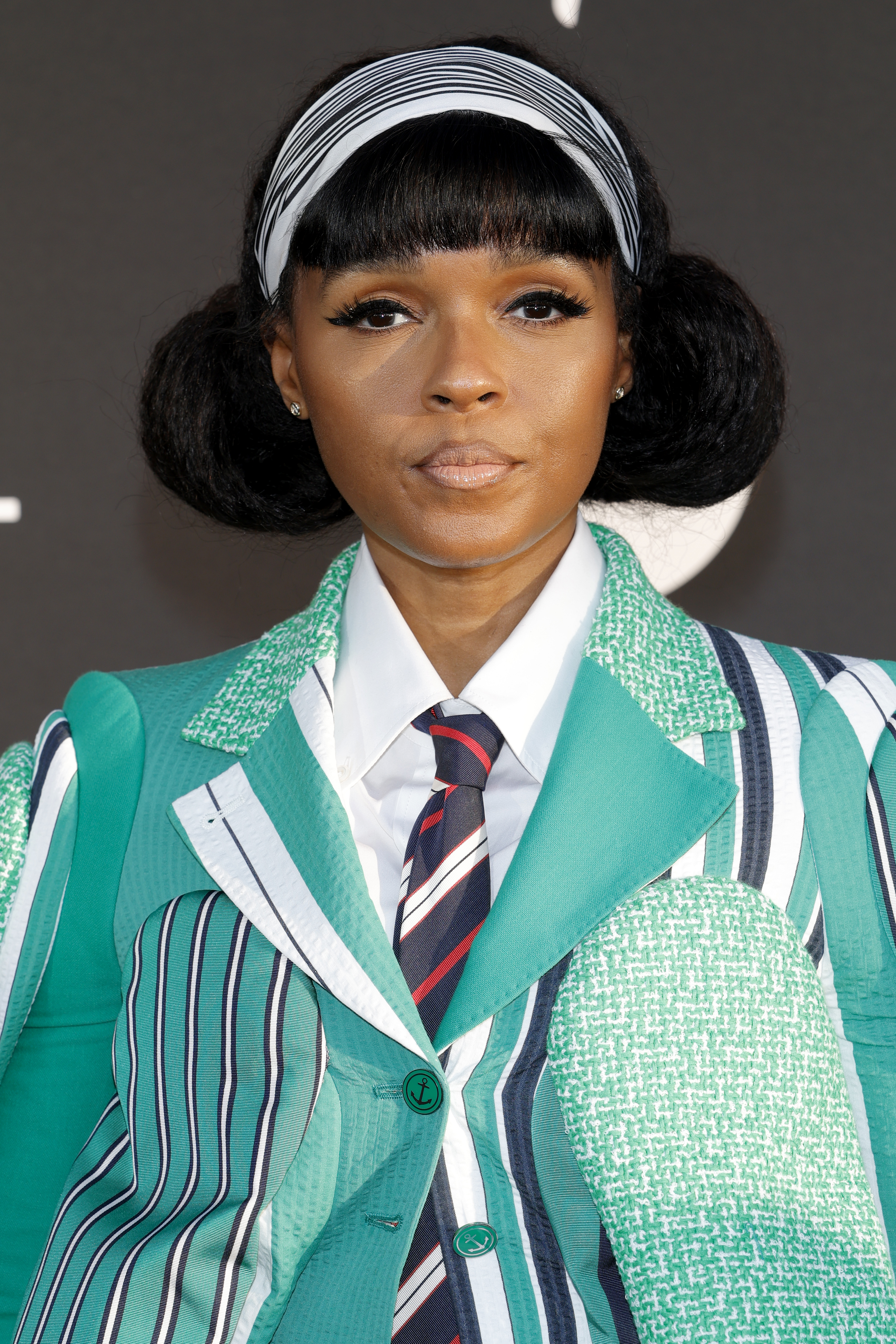 a black woman with dark hair pulled into two buns and blunt bangs wearing a black and white headband and a striped, green blazer. she is posing in front of a dark gray background