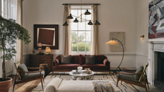 a large living room with a fireplace on the right, a set of two accent chairs, a small marble coffee table, a maroon sofa, a floor lamp, a large ceiling light, and a large window with curtains