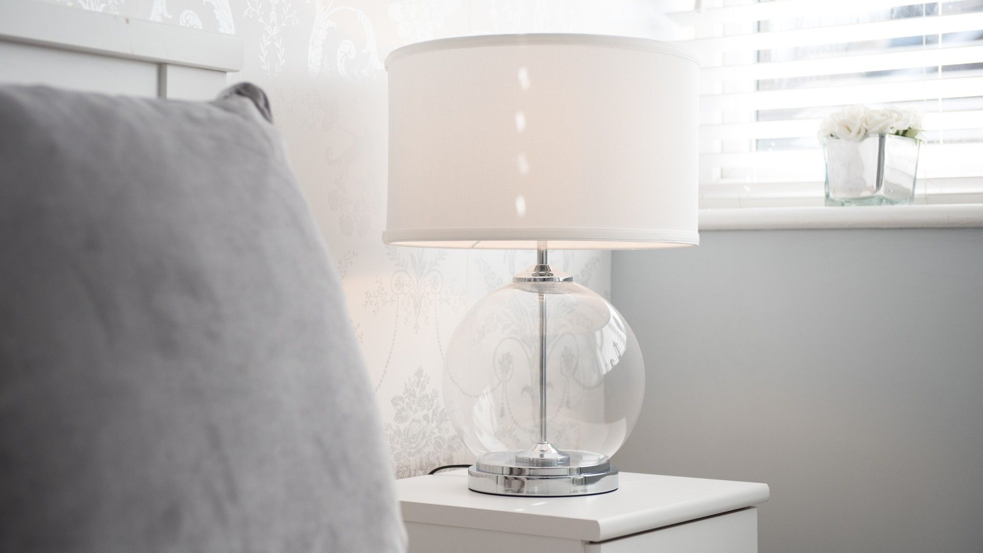 white lamp with glass base next to a gray bed in a light white bedroom