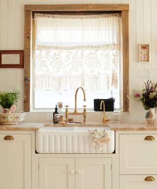 fluted butler kitchen sink undermounted in paneled cream kitsch kitchen with lace curtains, shaker cabinets, brass fixtures and orange marble counters