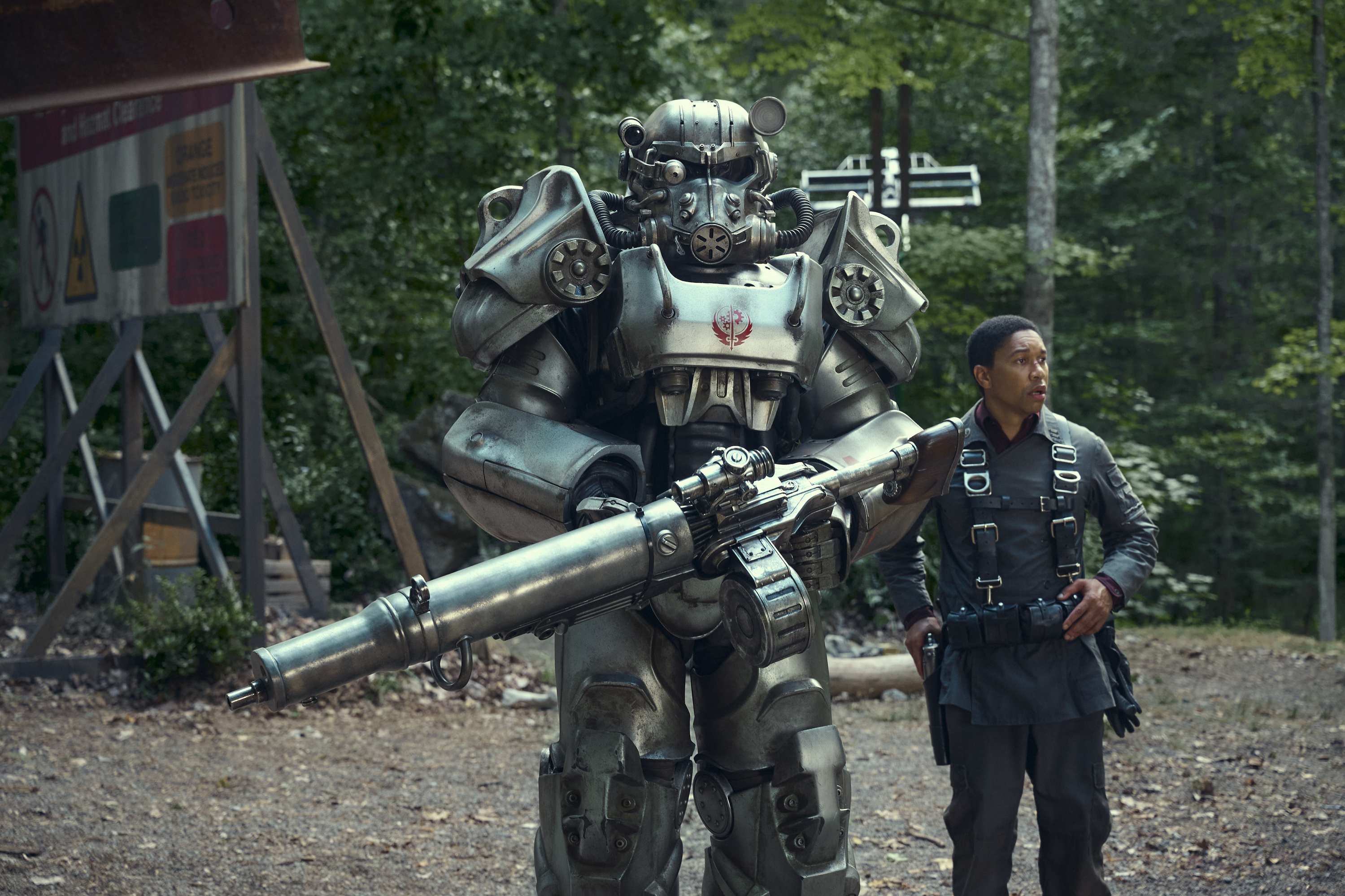 a man (aaron moten as maximus) in a jumpsuit stands to the right of a large mechanical suit of armor as they both stand in a forest clearing, in 'fallout'