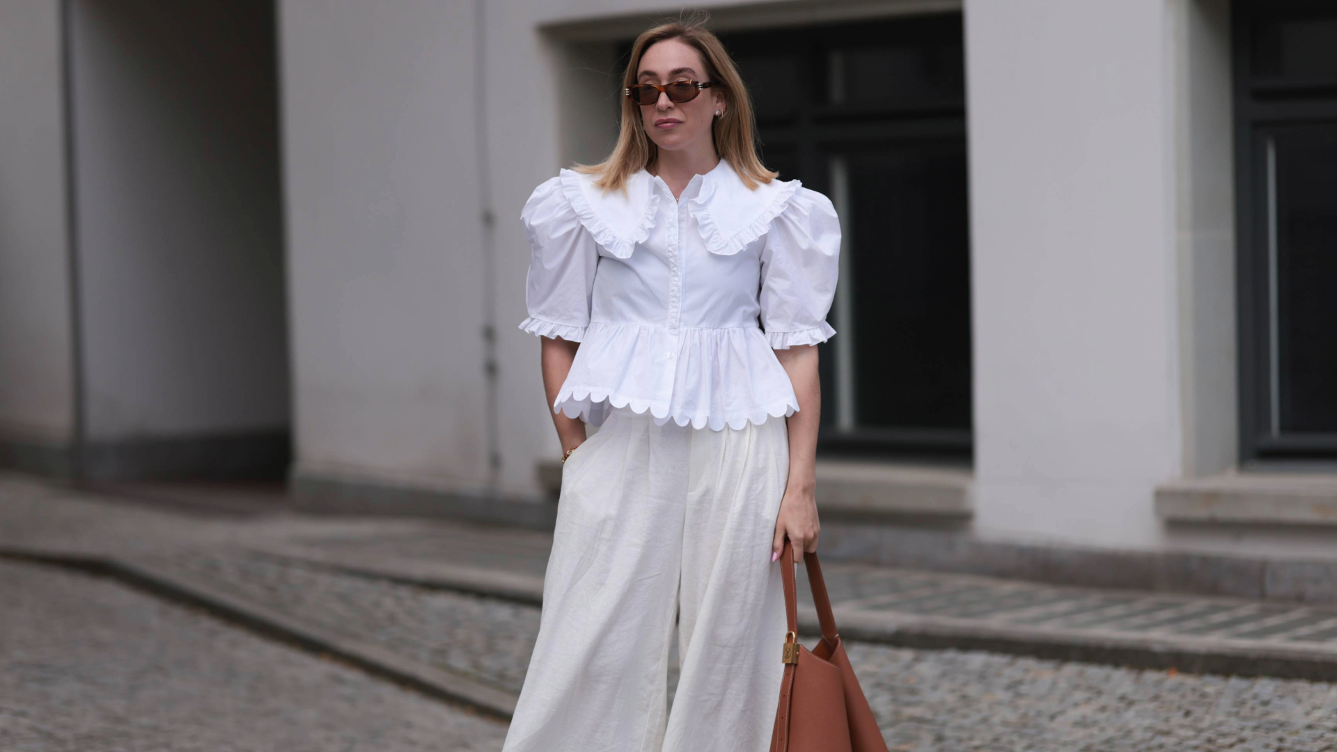 woman wearing white linen pants and a white embroidered top with a brown bag and brown shoes.