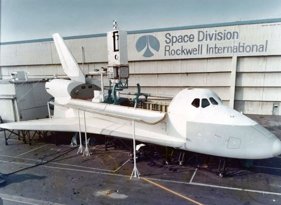 side view of a white space shuttle mockup, in front of a wall emblazoned with the words