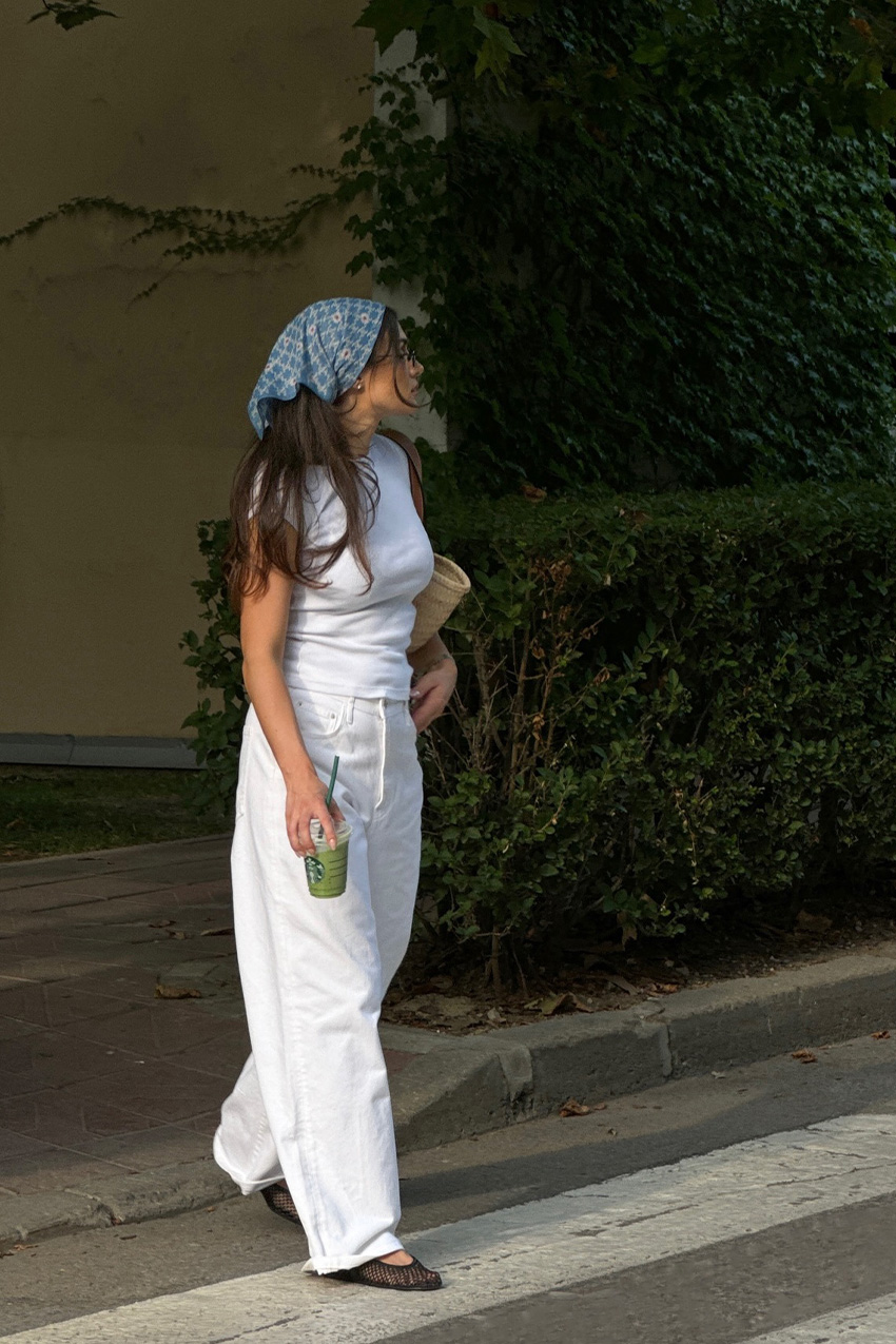 How to wear a bandana is shown in a photo of a woman walking a crosswalk wearing a blue printed headscarf with a white fitted t-shirt, white barrel leg jeans, black mesh ballet flats, and a raffia tote shoulder bag