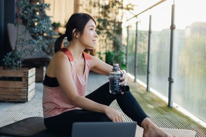 Fitness Christmas gifts: Young woman holding water bottle while sitting on exercise mat on the balcony