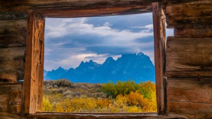 A window in an old log house looks out onto a beautiful mountain view.